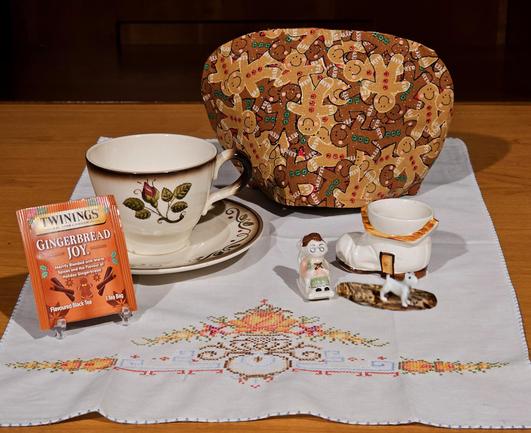Tea tableau upon an embroidered tea towel in autumn colors with a gingerbread man tea cozy and vintage Metlox china in ivory with brown edging and a single red rosebud design. There is a vintage ceramic figurine of a ruffled shoe with a door in it, a figurine of a bespectacled granny knitting in a rocking chair, and a tiny white terrier dog on a rug. The tea is Gingerbread Joy black tea from Twinings.