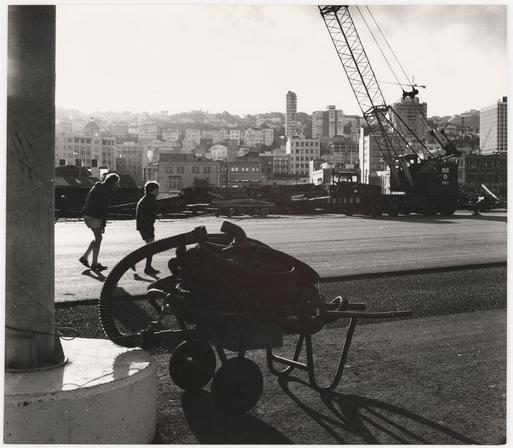 The black-and-white photograph captures an urban waterfront scene, likely a port area given the presence of cranes and construction equipment. In the foreground is a wheelbarrow filled with what appears to be construction materials or waste; it's tipped over on its side with various items spilling out onto the ground. The shadow cast by this structure suggests strong sunlight coming from above.

Two individuals are present in the scene, both moving away from the viewpoint of the camera and heading towards a distant area where another person is walking alone. Their attire and posture suggest casual movement rather than laborious work, which contrasts with the industrial setting around them.

In the background, there's an expansive view of buildings that could be indicative of a cityscape during daylight hours due to visible sunlight reflections off their surfaces. The overall atmosphere feels somewhat desolate except for these figures and hints at ongoing construction or maintenance activities in the vicinity.