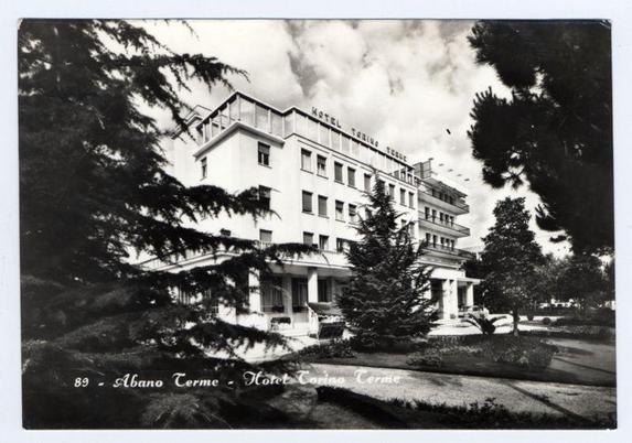 The black and white photograph depicts a multi-story building labeled "Hotel Torino Terme" in large, bold lettering across the top. The architecture suggests a mid-20th-century European design with symmetrical windows, balconies on some floors, and an overall clean facade. In front of the hotel is landscaped greenery including trees and manicured bushes, indicative of well-maintained grounds possibly for guests' leisure or relaxation. A pathway leads up to what seems like a main entrance area. The photograph's context appears vintage based on its style and quality; it likely dates from an earlier era given its monochrome presentation and the fashion implied by clothing visible in some windows. Below, text reads "89 - Abano Terme - Hotel Torino Terme," suggesting that this image is part of a series or collection related to the location known as Abano Terme, which could be associated with spa treatments considering such names typically relate to therapeutic mineral water baths and spas.