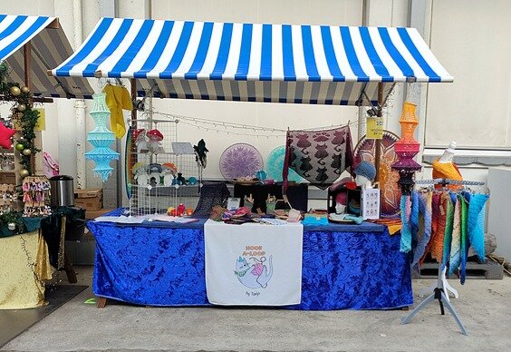 Market stall under a blue-and-white striped canopy, featuring handmade crochet items. A shiny blue tablecloth covers the table, which displays colorful hats, small decorative crochet pieces, and accessories. On the table stands a wire grid holding various crocheted Amigurumi figures. To the right, a rack presents an array of crocheted shawls. A sign reads “HOOK A LOOP by Tanja” and includes a cartoon cat holding a crochet hook and yarn.