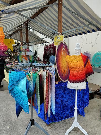 Outdoor market stall under a striped canopy, showcasing handmade crochet garments and accessories. A mannequin displays a gradient crochet shawl transitioning from yellow to red to purple. A rack to the side holds multiple crocheted shawls in varied colors and patterns. Behind the stall, circular mandala-style crochet pieces are mounted for display. The setup includes string lights.