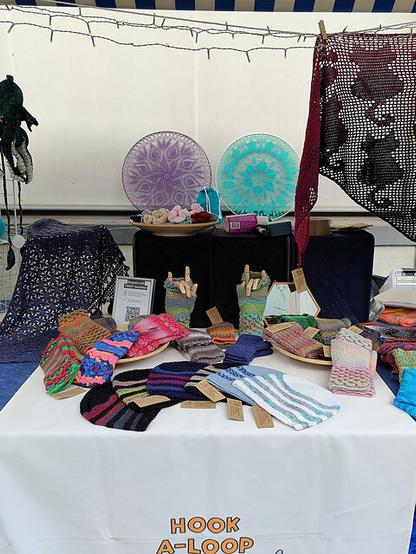 Market stall under a blue-and-white striped canopy, displaying handmade crochet items. A table is covered with colorful crocheted mittens, gloves, and small textile crafts, each labeled with tags. On the table, two large circular mandala-style crochet pieces in purple and turquoise are mounted upright. A burgundy crochet shawl hangs on the right side, and a dark blue crochet piece is draped on the left. String lights are strung across the top of the canopy. The tablecloth features the text “HOOK A LOOP” in orange and black.