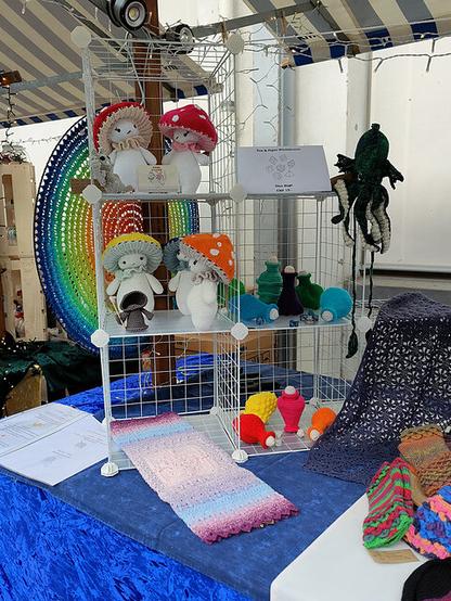 Craft fair display featuring handmade crochet items arranged on a white wire shelving unit with three levels. The top shelf holds crocheted dolls with mushroom hats in various colors and facial expressions. The middle shelf displays more mushroom-themed dolls, small crocheted potion bottles, and dice. The bottom shelf contains more potion bottles and dice. A gradient crochet piece in pink, blue, and purple is laid out on the table in front. To the right, additional crochet items include a dark blue piece with a star-like pattern. A crocheted Kraken hangs from the side of the shelving unit. Behind the display, a large circular rainbow-colored crochet mandala is mounted. The table is covered with a blue cloth