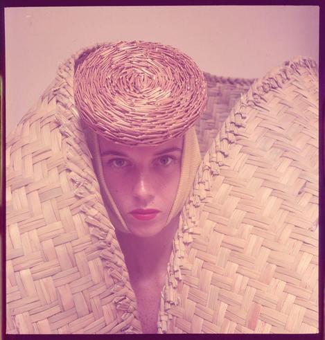 The photograph is of a person adorned with an intricately woven straw hat and what appears to be handcrafted, possibly bamboo or rattan, hood-like structures draped over their shoulders. The individual exhibits pale skin and striking blue eyes, accentuated by a vibrant pinkish-red lipstick.

In the background, there's no distinguishable scenery; instead, it seems that this image is intended for fashion rather than storytelling. It exudes an aura of sophistication with its vintage aesthetic, featuring soft yet warm tones that might suggest the use of sepia or light pinkish-purple hues to emphasize a classic and timeless quality.

The hat worn by the person adds another layer of elegance and uniqueness; it's not only stylish but also handcrafted which may hint at artisanal craftsmanship. The textures created through the weaving technique are visible, emphasizing an organic feel that resonates with natural elements like straw or bamboo used in its creation.

Considering additional information provided about Toni Frissell – a renowned photographer known for her work on Harper’s Bazaar and portraits of celebrities including Patsy Pulitzer - this image could be from one such collection. It's likely part of the 1940s-50s era, which is corroborated by its vintage look.

Moreover, given that Toni Frissell was renowned for capturing fashion moments with a blend of style and substance, it’s plausible to infer that this photo might  [...]