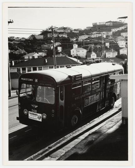 The photograph captures a vintage bus from an elevated angle, prominently featuring its front and side. The vehicle bears the registration number "308 TAUBMANS" on its license plate and is marked with several advertisements: one for "WELLEINGTON CITY TRANSPORT," another indicating it operates as No. 14 to Waidestown, and a third that reads "Transport." It's painted in dark tones, likely black or deep blue, which contrasts against the backdrop of suburban architecture consisting of houses on elevated terrains connected by roads and bridges. The scene is devoid of people, placing full emphasis on the bus as an element representing public transportation during its era. This image carries a historical snapshot quality to it, possibly dating back several decades due to the design elements present in both the vehicle and surrounding infrastructure.