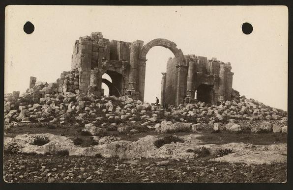 This black and white photograph depicts the ruins of a grand ancient structure, likely an archway or gate from what is now Jordan. The image captures only part of this impressive edifice, showcasing its weathered stone pillars that still stand amid heaps of rubble and rocky terrain. In one segment, there's a large, arched opening with significant dimensions, hinting at the building's historical significance as a monumental passageway or entrance.

In front of the ruins stands an individual in silhouette against the stark landscape, providing scale to this imposing structure. The sky is overcast but bright enough to reveal details on the stone and ground surface without harsh shadows. This image reflects the remnants of ancient architecture amid desolate surroundings, evoking a sense of historical exploration or archaeological study.

The caption indicates that additional information about this photograph can be found in 'East of the Jordan and Dead Sea' by Eric Matson and Edith Matson from around 1900 to 1920. The photo is credited to American Colony (Jerusalem) Photo Department, with a reference number for further context or research.

The URL provided directs viewers to an online archive where they can find more detailed information about this specific photograph by Eric and Edith Matson from the Matson Collection at LSE Library Special Collections: 'American Colony' photographic collection.