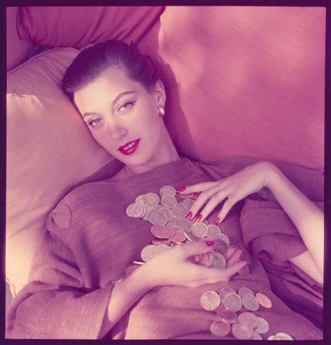 A color photograph of a woman reclining on a pink surface with her head resting against the armrest. She has dark brown hair, arched eyebrows and is wearing red lipstick. Her eyes are accentuated by eyeliner and she wears bright pink nail polish. The woman dons large earrings in addition to other jewelry adorning both hands which hold numerous coins that spill from a cupped palm towards her lap. Her fingers appear adorned with rings as well while the fabric of what appears to be an olive green sweater, is gathered around where it rests on top of several scattered silver and copper-colored coins.