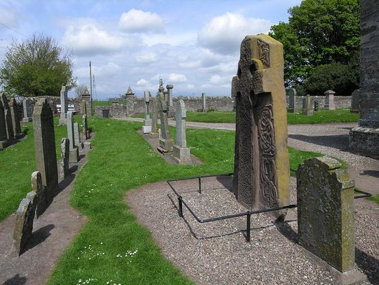 The Aberlemno Kirkyard Pictish cross slab. The image shows the kirkyard at Aberlemno in bright sunshine. A row of graves stands end on and starts in the bottom left of the frame, while another starts in the bottom right and converges with the first as it goes towards the wall on the far side of the kirkyard.  The second stone in the right-hand row, whose front is partly visible, is much larger than the other stones and carries a deeply carved cross and other carvings.