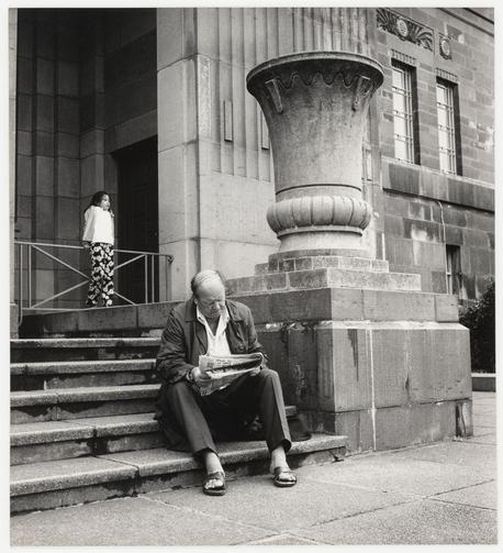 A black and white photograph depicting an older man seated on the steps of a classical building, reading what appears to be a newspaper. He is dressed in dark trousers, a light-colored shirt with rolled-up sleeves, a jacket open at the front revealing no tie or vest underneath, suggesting informality. His head is shaved except for short hair on top and he wears sandals, indicative of mild weather or casual atmosphere.

Next to him stands a younger woman leaning against an iron railing leading up to the entrance. She appears in profile view wearing dark trousers with large floral patterns down her calves and sleeves rolled over, revealing a white long-sleeved shirt underneath. Her hair is cropped close to the head but not shaved like the older man's.

The building features stone construction with two windows visible above the steps, one of which has an ornate frame. Prominently displayed beside them on the wall stands a large classical urn or pot atop what seems to be a pedestal, indicative of institutional significance perhaps relating to art and culture given its setting at "National Art Gallery" as suggested by additional context.

The photograph exudes a sense of quiet contemplation and suggests an older generation's engagement with contemporary life through media. The young woman’s presence adds contrast in terms of age and possibly societal roles or narratives being played out simultaneously within the same space, c [...]