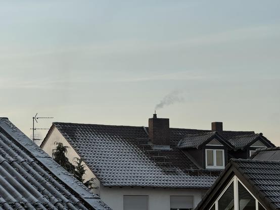 Ein Blick auf mehrere Dächer, die mit einer leichten Schneeschicht bedeckt sind, und einen Schornstein, aus dem eine Rauchwolke aufsteigt. Der Himmel ist blass, was auf einen kalten Tag hindeutet. Im Hintergrund ist eine Antenne zu sehen.