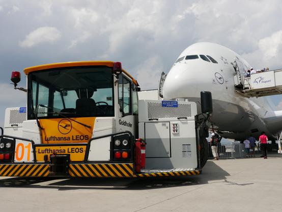 Aus der Froschperspektive ist die Nase eines Airbus A380 der Lufthansa zu sehen. Rechts und links sind direkt hinter dem Cockpit Zugangstreppen positioniert. Vor dem Flugzeug steht eine Zugmaschine, die Flugzeuge auf dem Boden manövriert, in weiß und Gelb lackiert mit dem Lufthansa-Logo und der Aufschrift "Lufthansa LEOS".