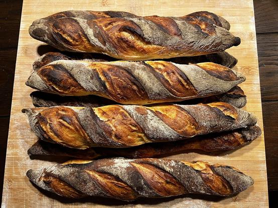 Auf einem dunklen Holztisch liegt ein Holzbrett. Darauf liegen zahlreiche frisch gebackene Brötchen mit rustikalem Ausbund.