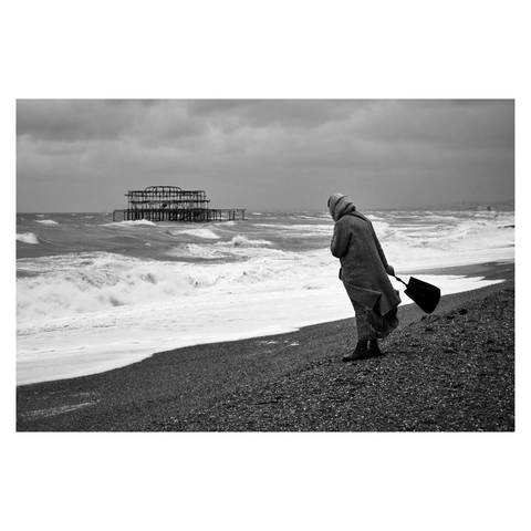 This is a landscape format black and white image of a woman standing on the pebbles of Brighton beach looking out across stormy waves. She is placed in the front right foreground. She wears a headscarf and clasps her coat at the neck. Her coat and a tote bag are caught by the wind coming off the sea. The burnt out West Pier is visible in the background to the left. Taken with a Sony RX100.