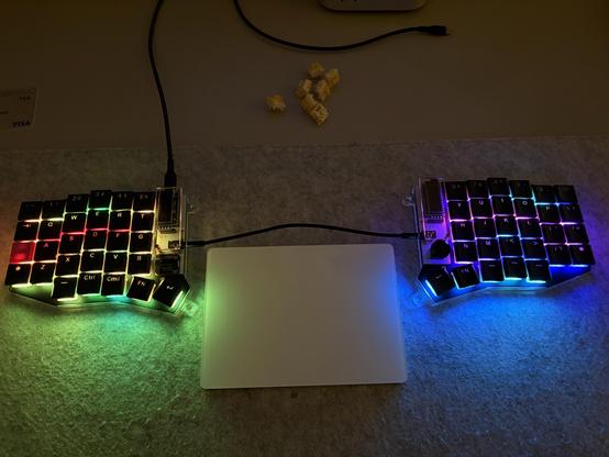 A photo of a colourfully lit split keyboard with a white computer trackpad between the halves. Yellow key switches are visible at the top of the photo.