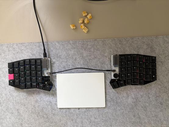 A photo of a split keyboard with a white computer trackpad between the halves. Yellow key switches are visible at the top of the photo. The board is on a grey desk mat.