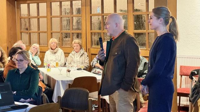 Mann mit Mikrofon spricht vor Publikum in einem hellen Raum mit Tischen und Stühlen. Eine Frau steht am Rand und schaut zu.