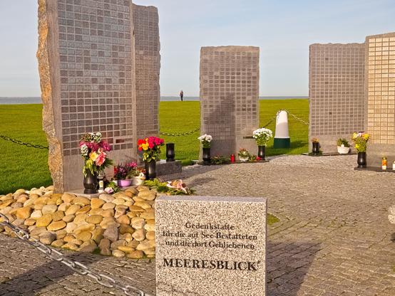 We are at the seaside, even if it is only faintly visible in the background. It is a sunny day. We are standing at a memorial for people who remained at sea. Either they were buried there or they perished. We see five tall, narrow blocks of stone or cement. Each of them has plaques with the names of the deceased. If you didn't know what you were looking at, you might think they were skyscrapers with windows reflecting the sunlight. In front of each stone block are vases with fresh, colourful flowers. In the foreground is a smaller stone. On it is written: "MEERESBLICK (sea view). Memorial to those buried at sea and those who remained there."