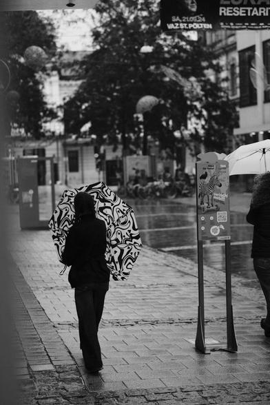 Das Foto zeigt eine Straßenszene in Schwarz-Weiß in der St. Pöltenter Innenstadt an einen regnerischen windigen Tag. Man sieht eine Person mit Regenschirm der durch den Wind verbogen ist.