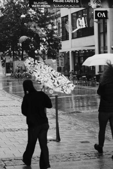 Das Foto zeigt eine Straßenszene in Schwarz-Weiß in der St. Pöltenter Innenstadt an einen regnerischen windigen Tag. Man sieht eine Person mit Regenschirm der durch den Wind verbogen ist.