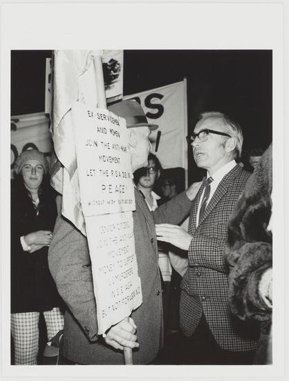 The black and white photograph depicts a group of individuals gathered, likely at a political rally or protest. A person is prominently holding up a sign with handwritten text that reads "EA-SERVAI-EMEN AND WOMEN JOIN THE ANTI-WAR MOVEMENT LET THE RASADERS PEACE" followed by smaller print ending in an obscured phrase and the word 'LOVE'. They are wearing glasses, a houndstooth patterned suit jacket over a light-colored shirt with a tie. Behind this person is another individual partially visible; they wear a dark coat or blazer.

The background includes several other people who appear to be part of the crowd. Some have their faces obscured by others in front of them and are difficult to discern individually due to partial visibility, but there's an impression that many more individuals were present at this event.

There is also another banner with visible text "VS" suggesting it could be a part of larger wording or logo not fully shown here, which may represent the name of an organization or group. The setting seems to be outdoors during nighttime as indicated by the darkness surrounding them and possibly artificial lighting on their faces from above.