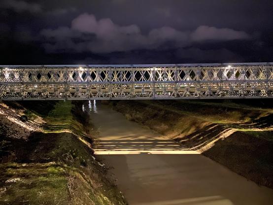 A metal bridge illuminated at night spans a waterway. The surrounding area features grassy banks and reflects the bridge's lights. Dark clouds are visible in the night sky.