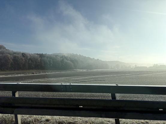 Eine frostige Landschaft, von einer niedrigen Sonne beschienen, Dunstschwaden in der Luft, gen Horizont in helles Weiß verlaufend. Im Vordergrund Leitplanken, das Foto scheint aus einem Auto aufgenommen.