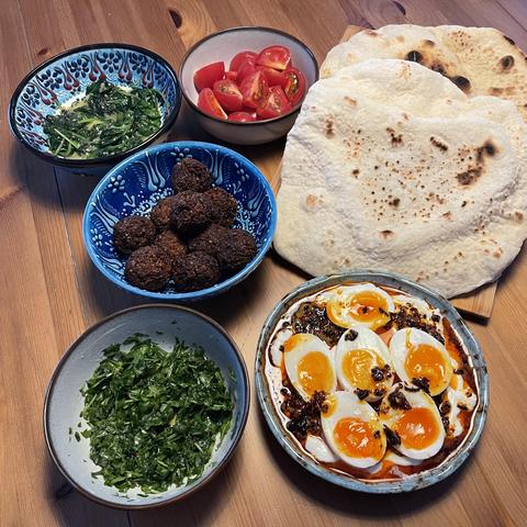 Na drewnianym stole stoi kilka misek: jedna z falafelami, druga z pomidorkami koktajlowymi, kolejna z duszonym szpinakiem oraz miseczka z posiekaną rukolą. Obok leżą świeże pity. W największej misce znajdują się jajka na półmiękko podane na jogurcie z ostrym masłem i przyprawami.