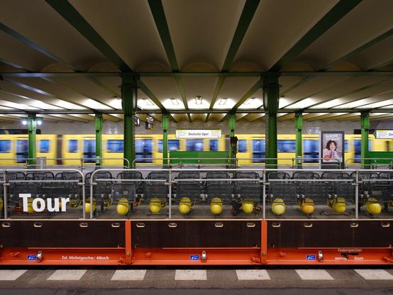 Das Foto zeigt den U-Bahnhof Deutsche Oper in Berlin quer über die Bahnsteige fotografiert. Im Hintergrund fährt ein U-Bahnzug - durch die Bewegung verschwommen abgebildet. Niemand steht auf dem Bahnsteig. Im Vordergrund steht ein offener Wagen mit Geländern und einfachen Sitzgestellen. Die Sitze stehen längs zur Fahrtrichtung und zeigen nach außen. Die Sitzflächen sind hochgeklappt. An jedem Sitz ist ein gelber Helm befestigt. Im Vordergrund ist die Randmarkierung des Bahnsteigs sichtbar. Wenige Farben bestimmen das Bild: das Rot des Untergestells der offenen Wagen, das Gelb der Helme und der U-Bahn sowie das Grün der Stahlträger und Deckenelemente. The photo shows the Deutsche Oper subway station in Berlin, photographed across the platforms. In the background, a subway train is moving—blurred by the motion. No one is standing on the platform. In the foreground is an open car with railings and simple seating frames. The seats are positioned lengthwise in the direction of travel and face outward. The seats are folded up. A yellow helmet is attached to each seat. The edge marking of the platform is visible in the foreground. Few colors dominate the image: the red of the undercarriage of the open cars, the yellow of the helmets and the subway, and the green of the steel girders and ceiling elements.