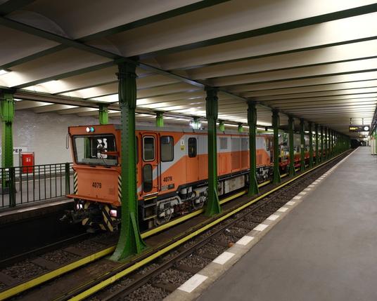 Das Foto zeigt eine orangefarbene Lokomotive mit angehängten offenen Wagen auf einem U-Bahnhof. The photo shows an orange locomotive with open cars attached at a subway station.