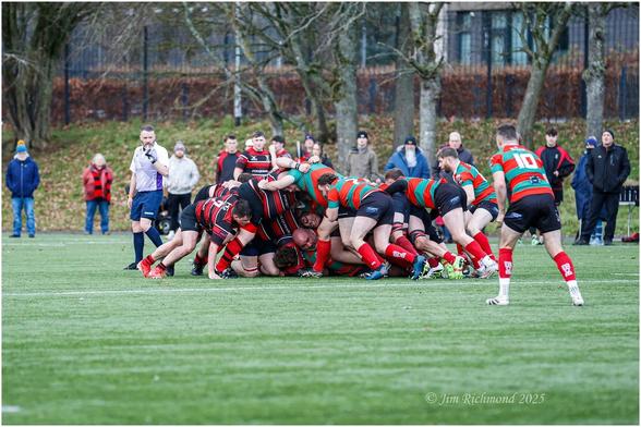 A rugby match scene with players in a collapsed scrum. The team in red and black stripes is engaged with a team in green and red stripes. A referee oversees the action, and spectators are lined up in the background.