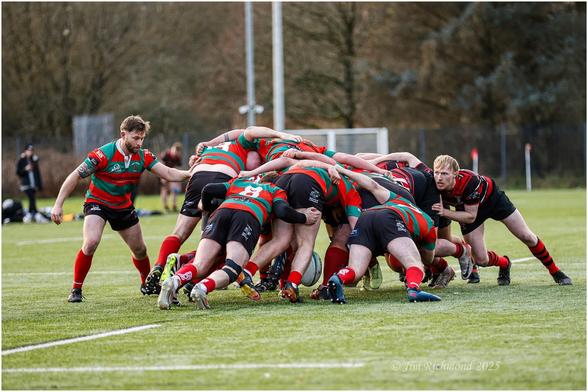 A rugby scrum involving players in red and green jerseys on a grass field. Some players are engaged in the scrum, while one player stands apart, observing. The background includes trees and field markings.