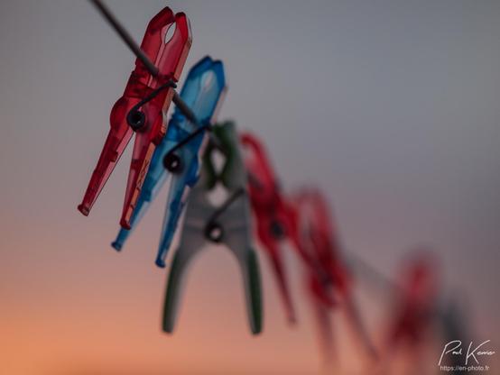 Photographie en couleur présentant quelques pinces à linge alignée sur un fil, captées avec une faible profondeur de champ, la première pince étant nette, la seconde un peu moins et déjà les suivantes sont nettement dans le flou. 
Le fil et les pinces forme, une diagonale du coin supérieur gauche au coin inférieur droit avec en arrière-plan, un ciel caractéristique de l'Arctique à cette époque de fin novembre, avec un soleil qui reste très bas sur l'horizon, passant directement de l'aube au crépuscule.