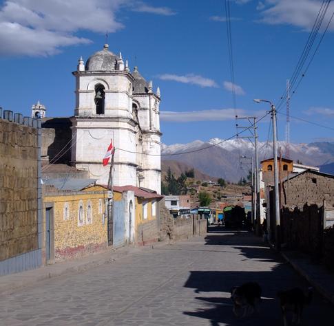Photograph of the main street in Cabanaconde, Peru. This is a village sitting just above the Colca Canyon and is the drop-off and exit point for visitors.
The photo is taken looking down the street, with the distinctive white twin-towered church on the left, each tower topped with a cupola. The buildings surrounding it are low and of the common concrete beam and block construction, with flat or low-pitched roofs. Looking straight down the street, in the distance are snow-capped mountains.
Power cables run overhead across the street, and a solitary red and white Peruvian flag is hoisted above a building on the left, in the foreground.