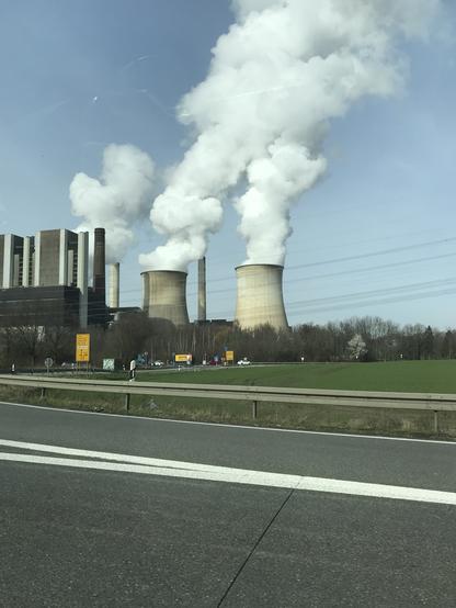 Zwei mächtige Kühltürme eines Braunkohlekraftwerks, aus denen eine Menge weißer Dampf in den blauen Himmel aufsteigt. Davor sind eine Autobahnabfahrt und Straßenschilder zu erkennen, rechts ein Stück grüner Acker. Stromleitungen ziehen sich durch das Bild.