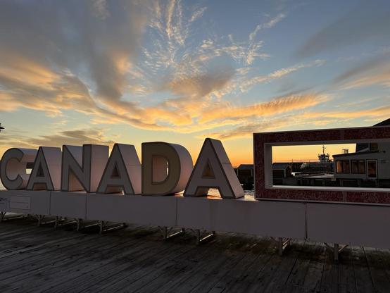 A large three-dimensional “CANADA” sign is backlit by a colourful sunrise on a wooden boardwalk. The dark brown boards run perpendicular to the sign, which is shiny and white with thin red outlines. To the right of the letters is a rectangular feature that frames a docked ferry. The sky has thin clouds emanating from the image center near the horizon, with the sky colours ranging from bright blue at the top to yellow to deep orange near the horizon. [Halifax (NS, Canada), November 2025]