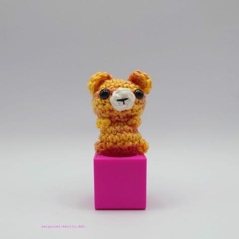 ein gold orangefarbener gehäkelter Bär auf einem pinkfarbenen Block a golden orange crocheted bear on a pink block