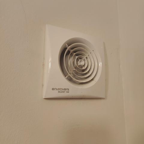An extractor fan in a bathroom wall with a circular grille that's permanently open.