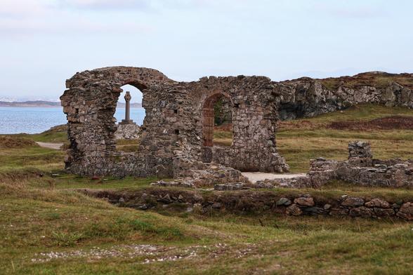 A picturesque scene of ancient stone ruins, likely the remains of a small chapel or church, perched on a grassy hillside overlooking the sea. The ruins feature two prominent arched windows, their weathered stone frames standing against the backdrop of a calm, expansive body of water. In the centre of the ruins, a tall, intricately carved Celtic cross stands as a focal point, adding historical and cultural significance to the scene. The surrounding landscape is a mix of grassy patches, rocky terrain, and low-lying stone walls, extending towards the coastline. The sky above is overcast, casting a soft, muted light over the tranquil setting. The overall atmosphere evokes a sense of timelessness and serenity, blending natural beauty with historical charm.