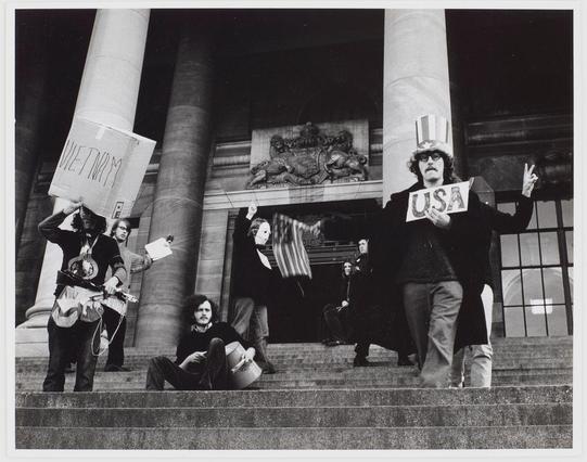 The photograph depicts a group of protesters gathered outside what appears to be an official or governmental building, with large pillars and grand architectural features. In the foreground on the left side, two individuals are standing; one is holding up a sign labeled "VIET NAM" while the other has their arm outstretched in a motion suggesting throwing something into the air. Another person sits down behind them playing an acoustic guitar.

In the center of the image stands another individual wearing glasses and a top hat with stripes resembling those on the American flag, holding up signs that read "USA." They are making hand gestures often associated with peace or victory—a raised index finger and two fingers forming what resembles the number 'V' sign. In the background, several other people can be seen sitting on steps; one is lying down while another sits casually.

The scene conveys a sense of protest against war, possibly related to Vietnam given the signs present in the image. The overall atmosphere appears peaceful yet assertive as demonstrators voice their opinions and engage with passersby or authorities within sightlines of what seems like an important building setting.