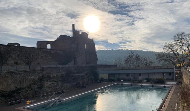 Im Vordergrund das Schwimmbecken eines Freibads. Das Wasser ist nicht abgelassen und obenauf gefroren. Links Teile einer Burgruine, darüber in einem leicht bewölkten Himmel die hell strahlende Sonne. Rechts nackte blattlose Bäume.

In the foreground, the swimming pool of an outdoor pool. The water is not drained and has frozen on top. On the left, parts of a castle ruin, above it in a slightly cloudy sky, the bright shining sun. On the right, bare leafless trees.