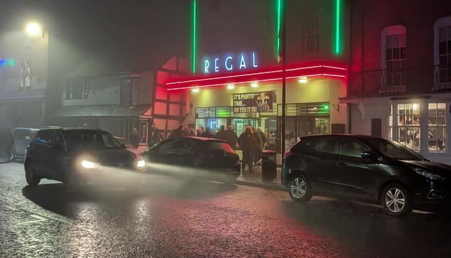 A somewhat foggy shot of the outside of the venue, a gorgeous 1937 facade of the theatre/cinema with red and green neon lighting.
People are exiting the building as a car drives past its headlights shining in the mist.