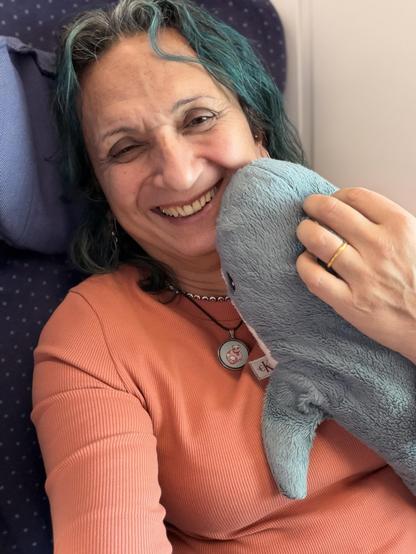 A smiling person with turquoise-streaked hair is sitting on a train, embracing a plush blue shark. They're wearing an orange long-sleeved shirt and a necklace with a cat pendant. Their left hand is holding the shark, and they have a gold ring on their finger.