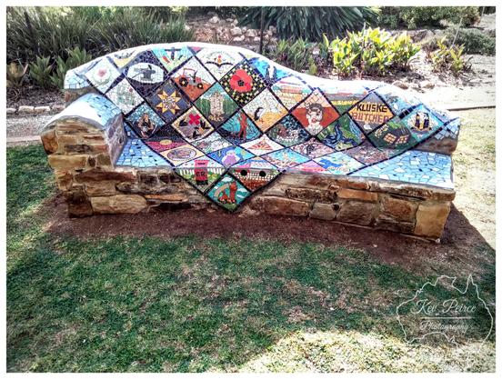 A unique outdoor stone bench, with the seat and backrest completely covered in a colorful mosaic.  The mosaic is arranged in a diamond or quilt pattern, with each tile depicting a different image, seemingly related to local history, trades, or farming, including a tractor, a poppy, an anvil, a pair of boots, and one section with the words "KLUSKE BUTCHER."  The bench sits on a patch of grass in front of a garden bed with lush green and brown foliage.