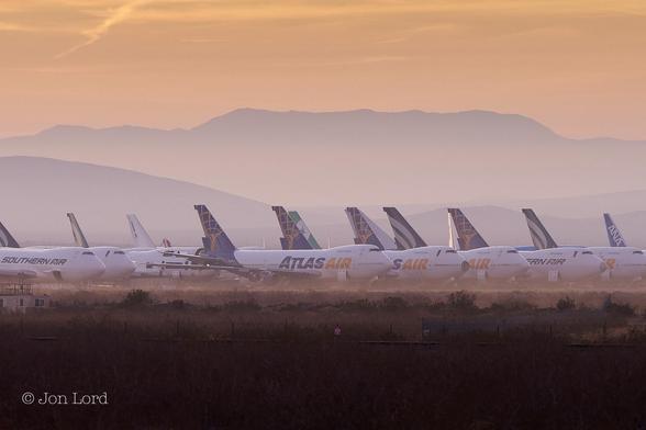 This is a colour landscape photo in landscape format of an aircraft graveyard in the desert at sunrise. Mojave, California (2016).

Stretching away from the camera is a wide expanse of desert scrub, sparse yellow grass and bushes, deep in shadow close to the camera and becoming more visible with increasing distance. A little way ahead is a tall fence running across the image from left to right, again in shadow. A third of the way up in the photo is a mass of jet airliners covering the entire width of the image. All in longterm storage and probably destined to be recycled into beer cans or some other high tech aluminum product. Most of the aircraft are Boeing 747 jets, ten or twelve in view, all with their airline's liveries and logos still painted on the planes. Atlas Air and Southern Air being in the majority. All the planes are facing noses to the right and parked in neat rows and lines. Beyond this aircraft graveyard and in the distance is a hazy and orange view of the San Gabriel Mountains lit by the still low rising sun.