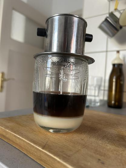 A clear glass jar sits on a wooden cutting board with a metal coffee filter apparatus on top. The jar contains a dark brown liquid with a lighter, creamy layer at the bottom, indicating separation of liquids. The metal filter has several protruding knobs. The background includes a tiled wall, various clear glasses, and a dark brown glass bottle with a pump dispenser. The overall scene appears to be in a kitchen setting.