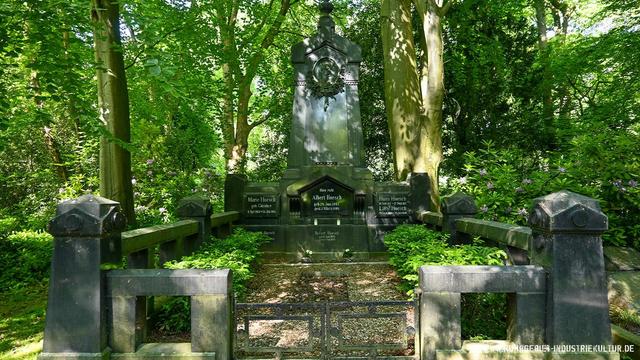 Monumentales dunkles Grabmal der Familie Hoesch auf einem grünen Friedhof. Hoher Grabstein mit Reliefmedaillon und mehreren Namensinschriften, umgeben von symmetrischen Steinbänken. Sonnenlicht fällt durch die Baumkronen.