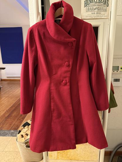 Foto eines roten Wollmantels, der auf einem Kleiderbügel vor einem Garderobenspiegel hängt. Der Mantel ist ungefähr knielang, tailliert und asymmetrisch mit drei Knöpfen geschlossen. Er hat einen halsnahen hohen Schalkragen. Die Knöpfe sind mit Mantelstoff überzogen.