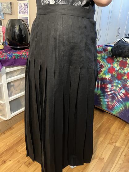 Front view of a black linen pleated maxi skirt taken in a room with hardwood fliers and printed tablecloths
