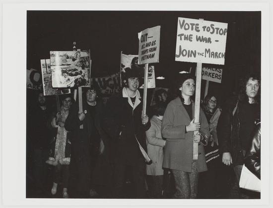 The photograph depicts a group of individuals participating in an anti-Vietnam War protest. They are holding signs with messages such as "Vote to stop the war – Join the March" and "Out now all U.S. troops from Vietnam." The crowd appears engaged, many looking towards something outside the frame, possibly speakers or focal points for their demonstration.

The people in the photo exhibit a range of emotions; some look serious, while others seem more contemplative. They are dressed warmly, suggesting it might be cold weather, and various age groups can be seen among them, indicating widespread public support for this cause.

In terms of composition, black-and-white tones dominate the image, which creates a somber atmosphere fitting the gravity of their message. The photo captures a sense of unity in purpose as well as diversity within those who oppose the war. There are no explicit indicators of specific locations or time periods beyond these general observations.