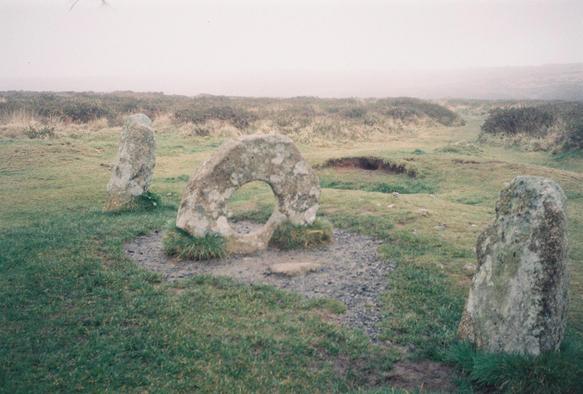 A grainy 35mm colour film photo of the Men-an-Tol, in penwith in Cornwall. It consists of a donut-shaped holed stone in the centre, large enough for someone to crawl through the middle of, and two small upright stones in front & behind the holed stone, forming a line. It lives on a moor which is green and brown in autumn, and the sky is grey and uniformly cloudy.